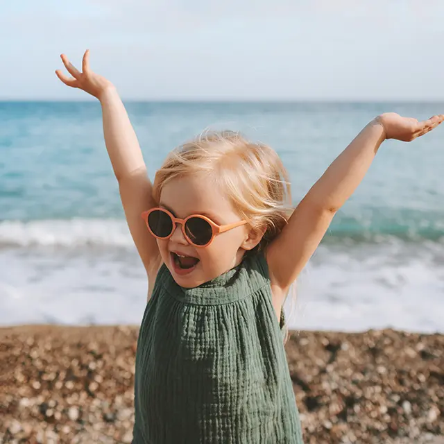 Ein kleines Mädchen mit Sonnenbrille am Strand streckt fröhlich die Hände in die Luft